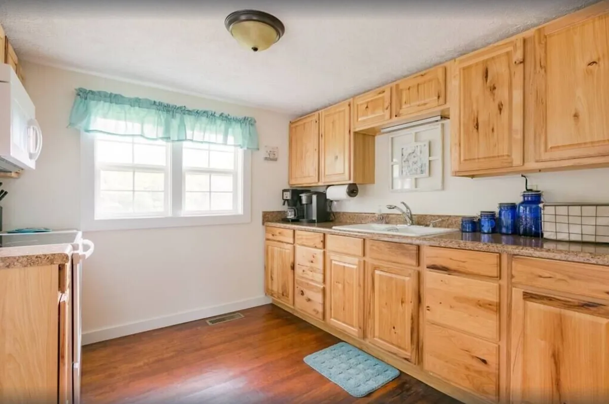 A cozy, warmly lit kitchen with wood cabinetry, blue accents, and double windows letting in natural light. The space feels inviting, perfect for gathering with family or enjoying a morning coffee. The kitchen is outfitted for cooking and socializing.