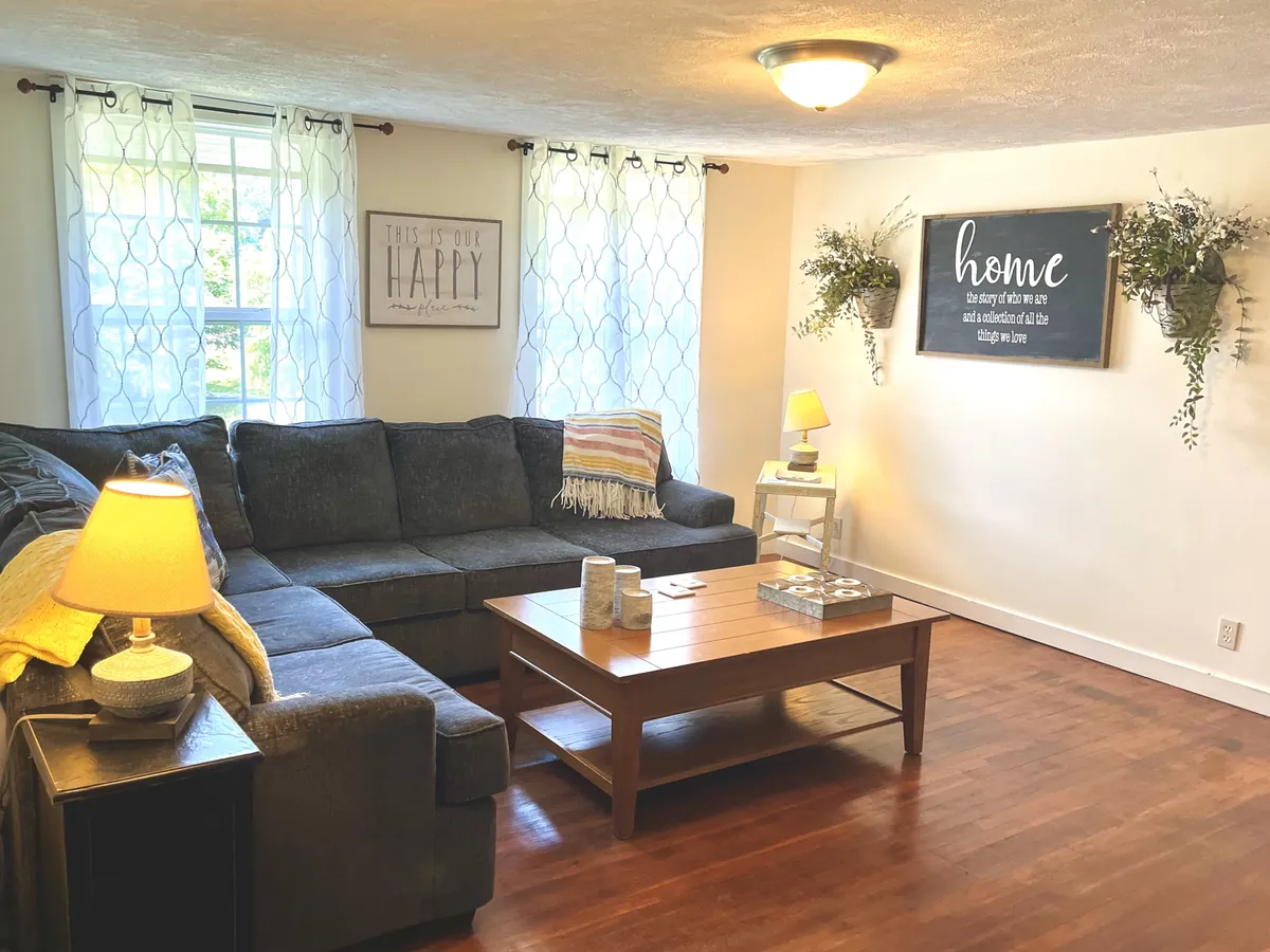 This image features a spacious living area with a large, inviting sectional sofa arranged around a coffee table. Decorative wall art and a soft lamp contribute to the cozy, welcoming atmosphere. The layout is open and perfect for gatherings, reading, or relaxing. The warm lighting and soft textures emphasize the comfort of the room.