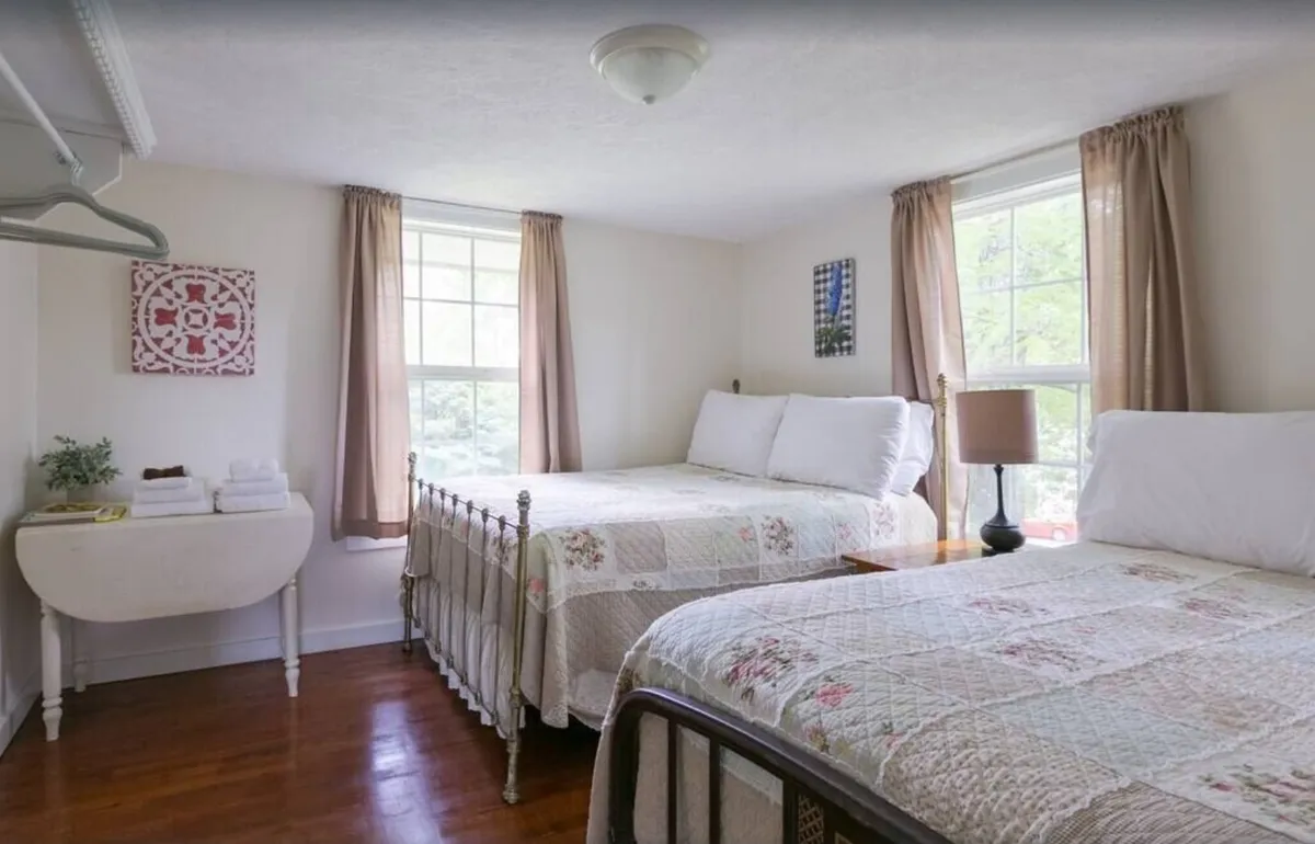 A bright and welcoming bedroom with two beds adorned in floral quilts, large windows letting in abundant natural light, wood floors, a small bedside lamp, and decorative wall art. The setting is inviting for families or friends to share.