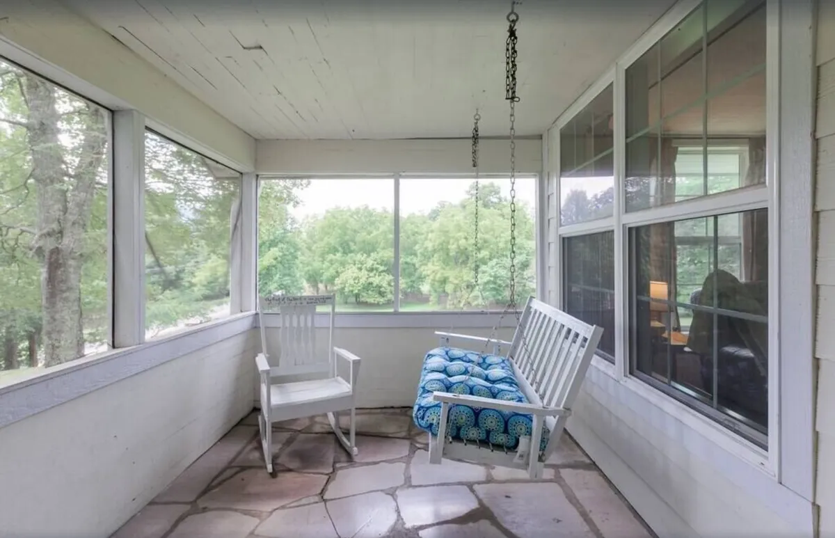 A peaceful sunroom featuring a swing bench with a blue patterned cushion, a white rocking chair, and large windows that bring in plenty of natural light and offer tranquil green views outside. The setting is ideal for relaxing with a book, sipping morning coffee, or simply enjoying the surrounding nature.