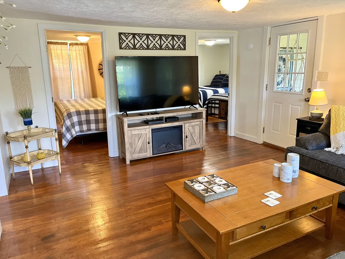 This image shows another angle of the living room, focusing on a comfy couch facing a television, with a coffee table and the entrance to a bedroom in view. Natural light bathes the space, reinforcing an open, airy feel. The scene is set for movie nights or peaceful mornings, with an atmosphere that feels both homey and versatile.