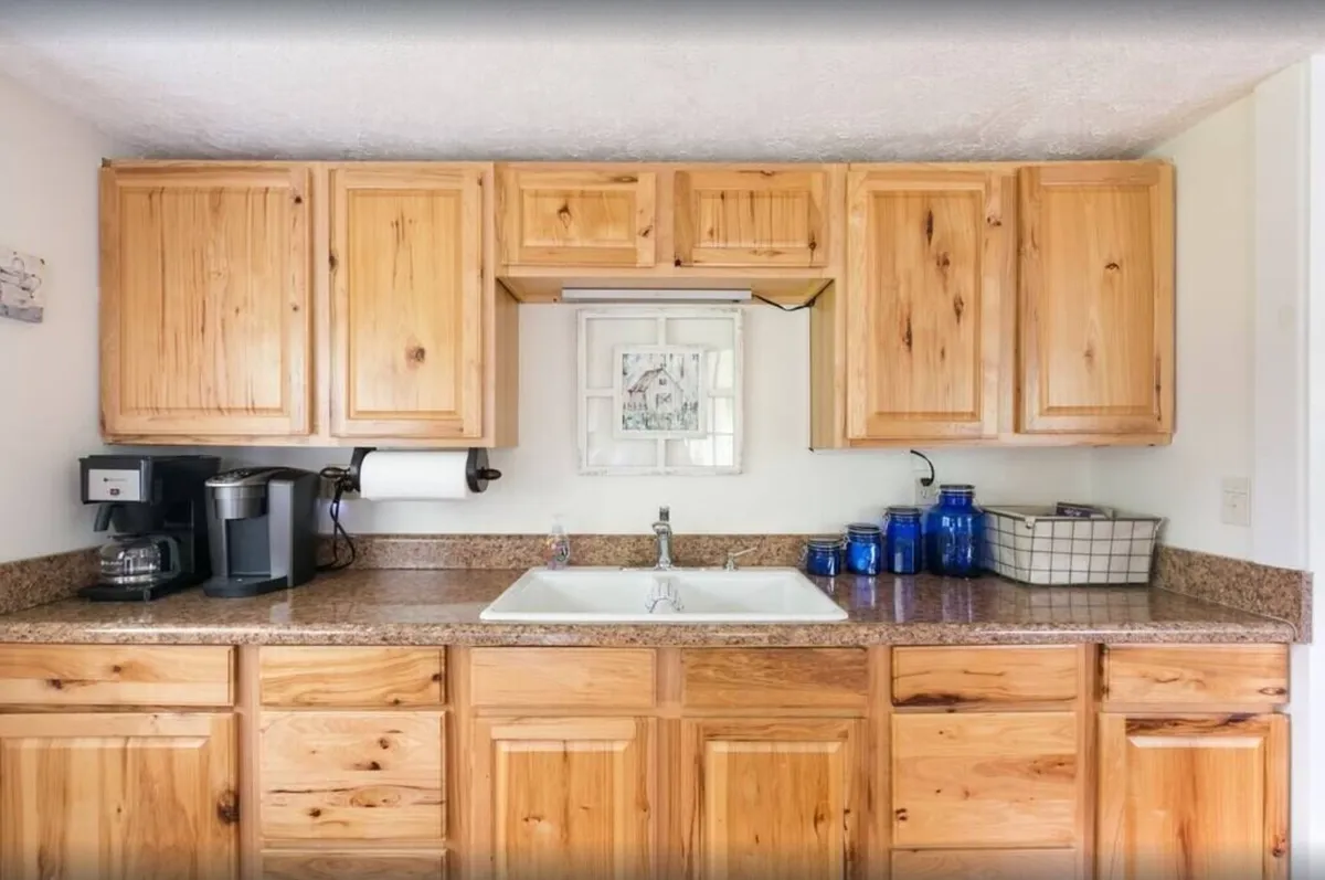 A homey kitchen scene featuring natural wood cabinetry, a granite countertop, blue glass jars, and a prominent sink area. The space is arranged for meal prep and conversation, suggesting easy socializing and shared experiences.