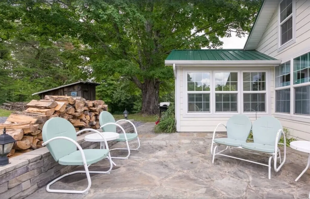 This exterior image showcases a tranquil outdoor scene featuring a wood pile, inviting outdoor seating, a large tree, and a structure with prominent windows. The space exudes a sense of relaxation and togetherness, with ample room for socializing and unwinding in nature.