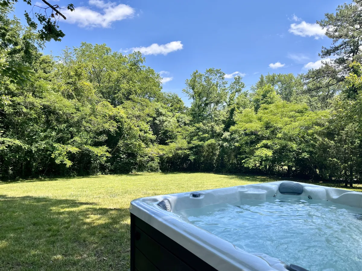 A wide exterior shot featuring the inviting hot tub nestled among lush greenery, open lawn space, and clear views toward the mountains. The scene feels tranquil and provides a sense of spaciousness, with ample room for pets to roam and guests to enjoy nature.