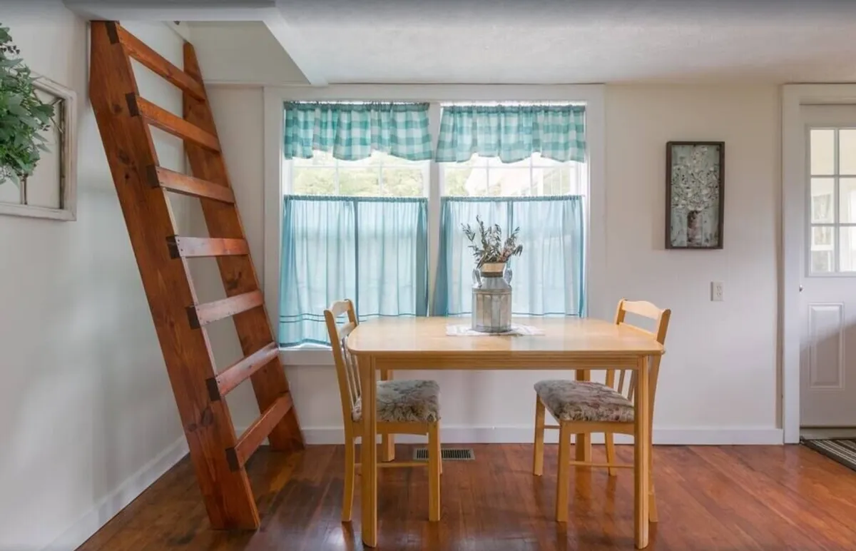 A cozy interior dining space with a rustic wooden ladder, a family-sized dining table, floral centerpiece, and soft curtains. The scene is warm and welcoming, ideal for shared meals, casual gatherings, or game nights after a day spent exploring local attractions.