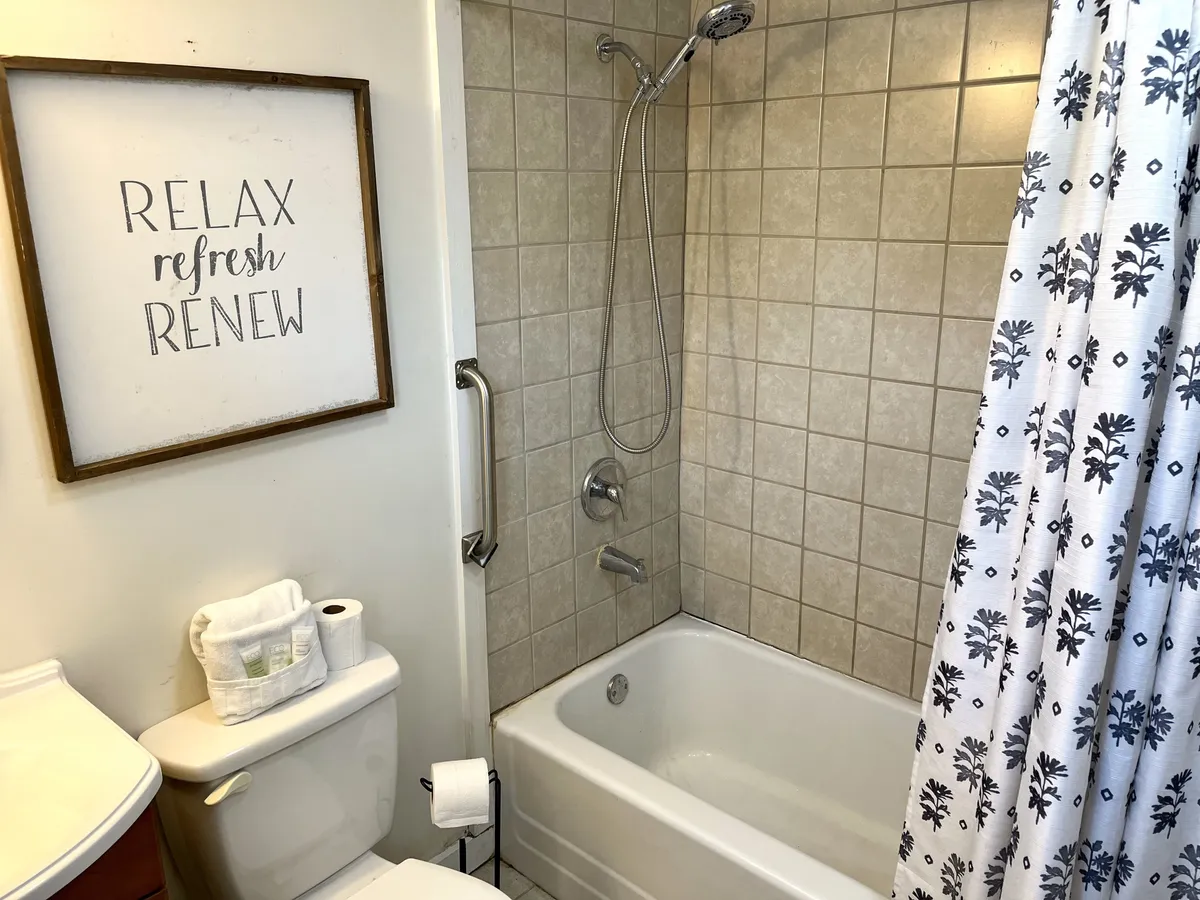 A modern bathroom with a combination bathtub and shower, a toilet, plush towels, and a framed decorative sign. The space feels clean, functional, and ready to support guests’ routines.