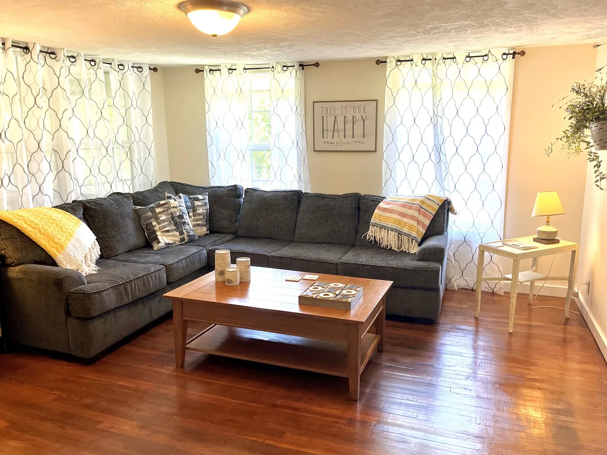 Captured from a slightly different angle, this image spotlights a large sectional sofa with decorative pillows, a wooden coffee table, and a window with curtains. Wall art that reads 'THIS IS OUR HAPPY' adds a personal, inviting touch. The overall mood is cozy and lively, suggesting a space designed for laughter and shared experiences.