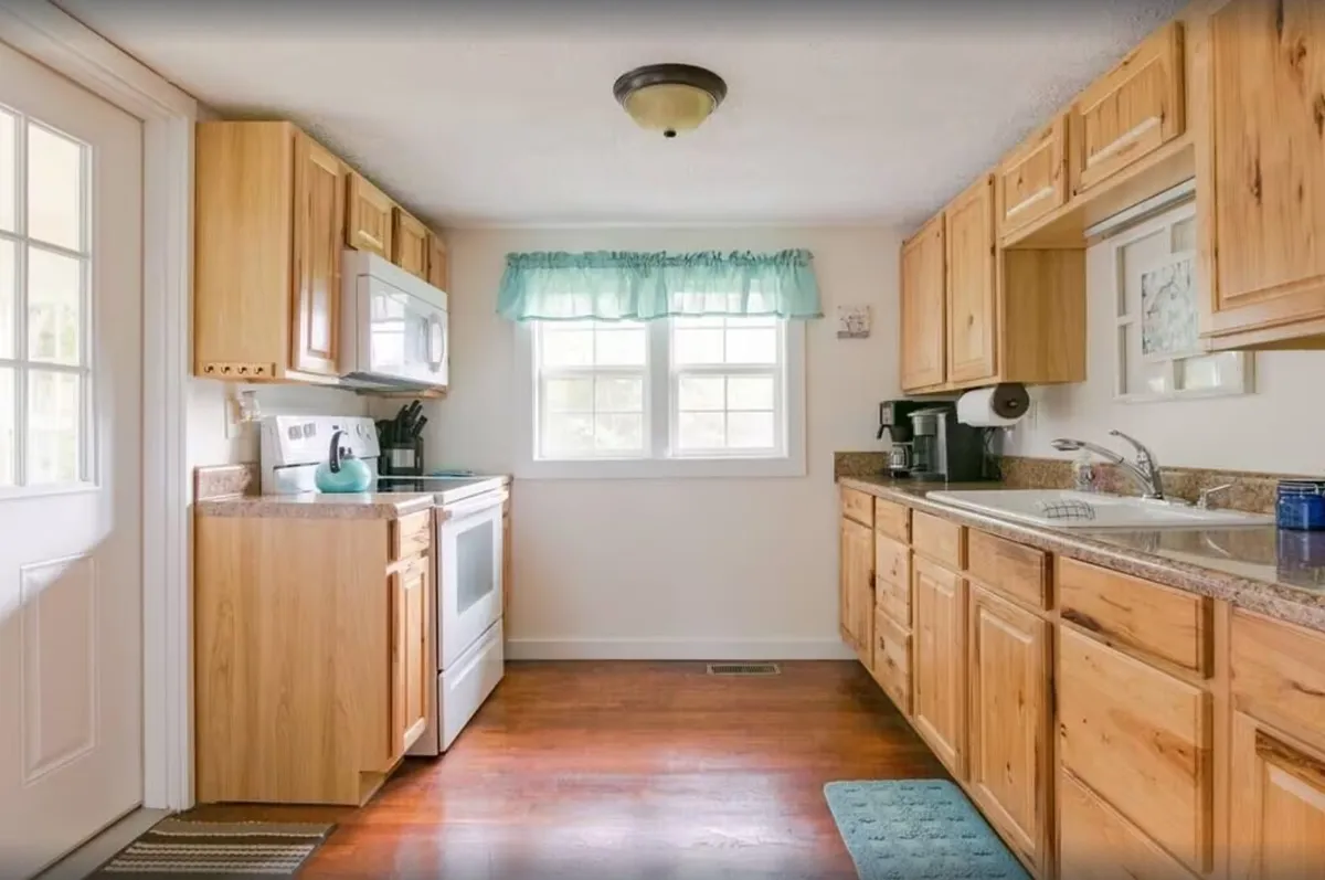 This image focuses on wooden cabinetry, white appliances, and a kitchen sink beneath a window with light curtains. The setup suggests a well-equipped, functional kitchen ready for family meals or group cooking sessions.