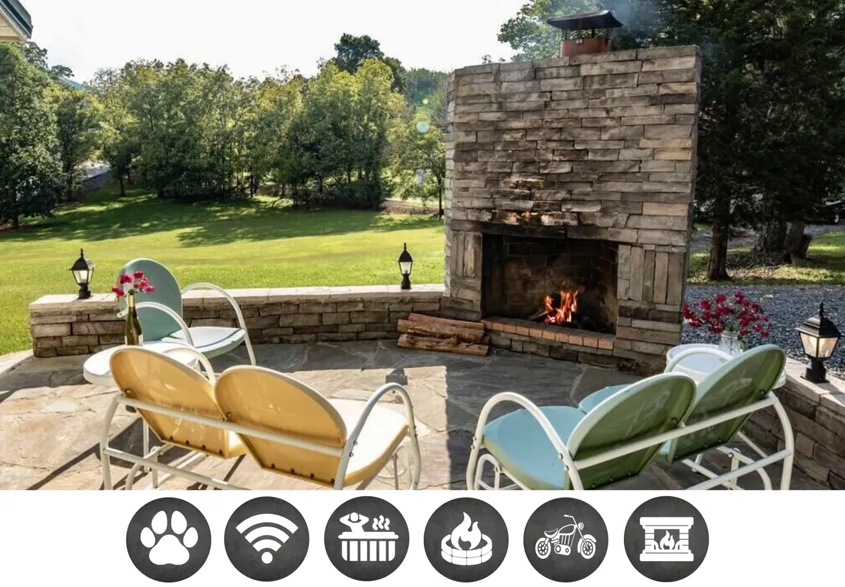 The image showcases a cozy outdoor seating area centered around a rustic stone fireplace, surrounded by lush greenery and softly glowing lanterns. The scene is set at sunset, with warm firelight reflecting off the stone and inviting guests to unwind and savor tranquil mountain evenings.