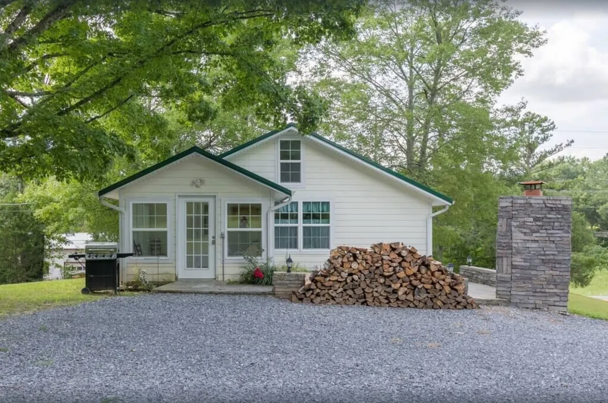 A cozy exterior scene featuring the house, a neat woodpile, and a prominent stone chimney. The outdoor space is primed for gathering, with areas suited for barbecuing or enjoying the warmth from the fireplace, surrounded by the tranquility of the yard and natural landscape.